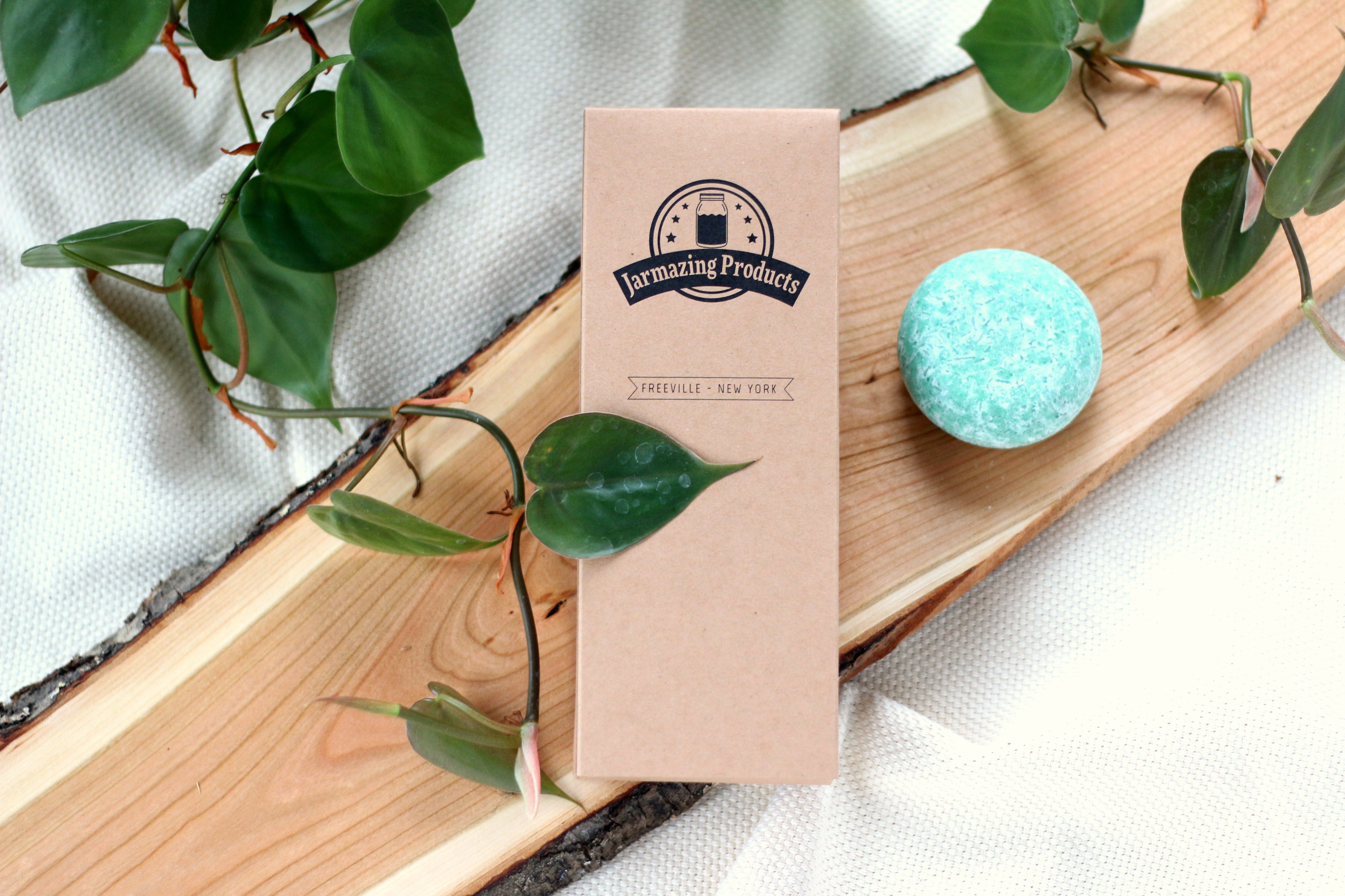 A bar of soap and a Jarmazing Products retail cardboard box on a decorative board, with green leaves streaming through.