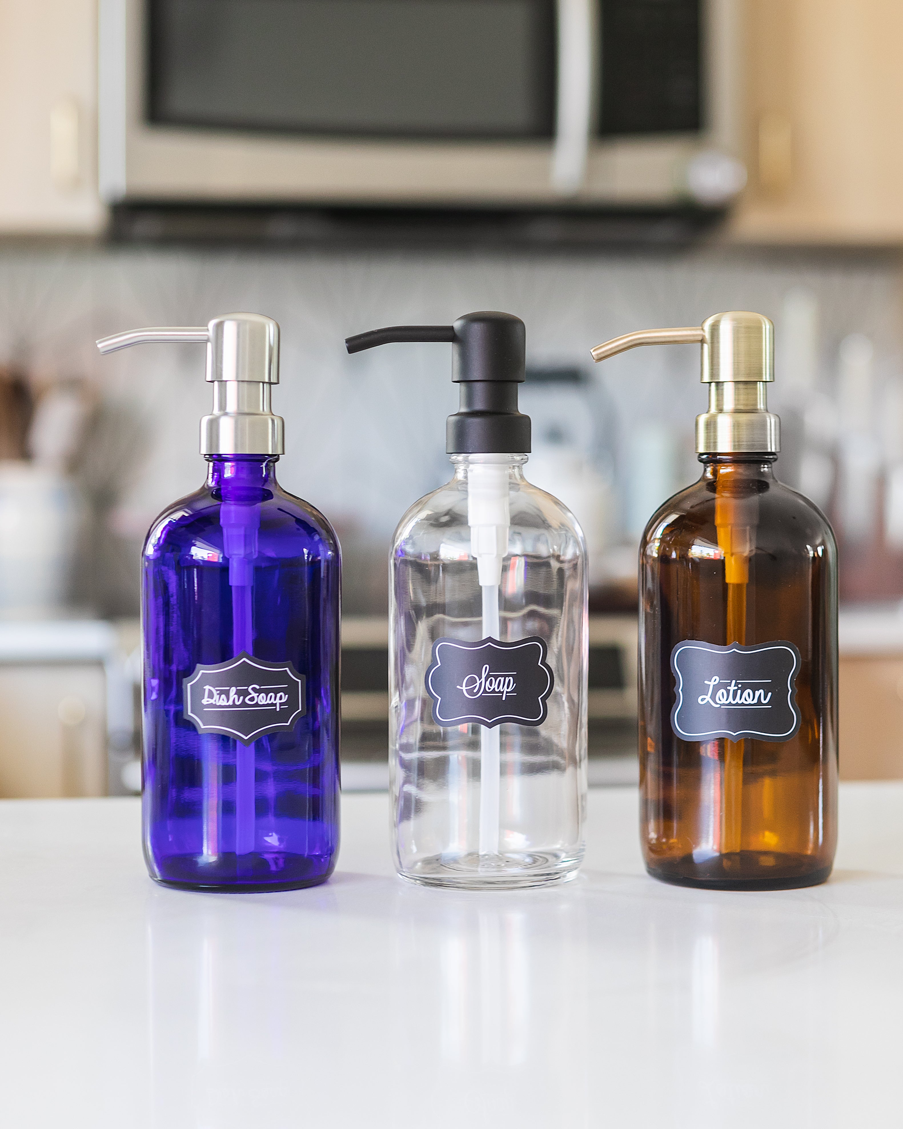 Set of three boston round bottles in blue, clear and amber glass, each with a bird-head soap dispense in stainless steel, black, or brass color, and each with a sticker stating dish soap, soap, or lotion.