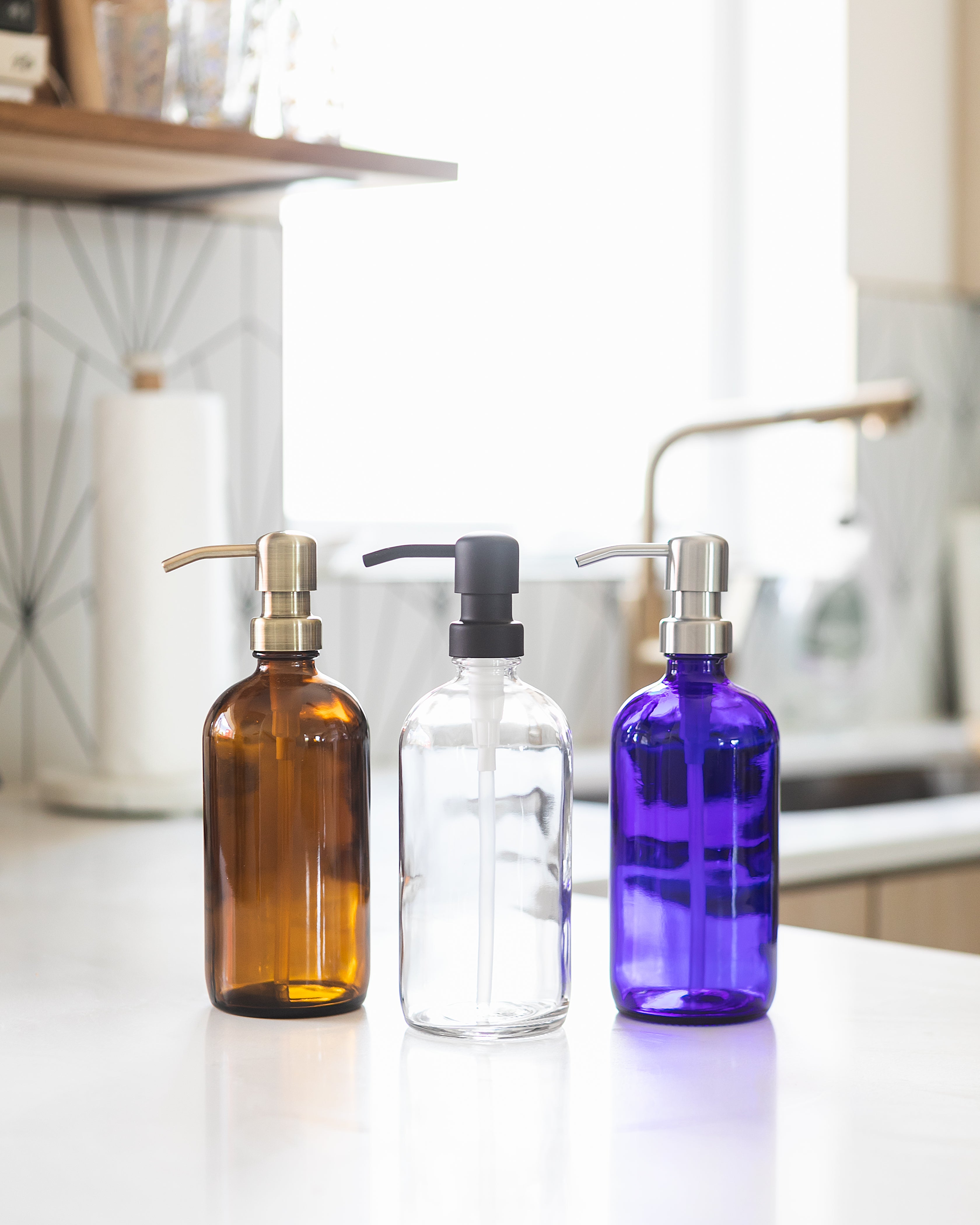 Set of three boston round bottles in blue, clear and amber glass, each with a bird-head soap dispense in stainless steel, black, or brass color